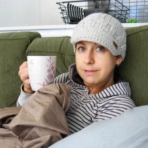 A woman in a grey hat sitting on a sofa with a coffee mug smirking at the camera